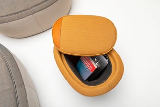 Gele poef met open compartiment waarin een zwarte doos en rode verpakking liggen, naast grijze zitmeubels.