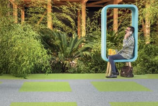 Man in geruite overhemd zit in turquoise belcel met houten poten, telefonerend. Achtergrond met groene planten en bamboe.