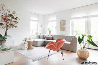 Lichte woonkamer met grijze hoekbank, roze fauteuil, witte vloerlamp, houten tafel, witte rug, planten en macramé wanddecorat