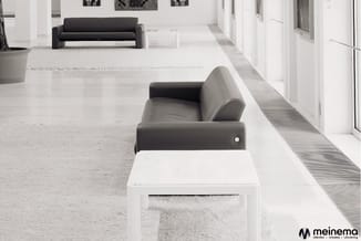 Moderne kantoorruimte met zwarte design fauteuils, witte ottoman, grijze tapijten en lichte houten vloer. Meinema-logo rechts