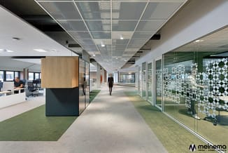 Modern kantoorinterieur met glaswanden, houten kolom, groene tapijtzones, mesh plafond en verlichte werkplekken met personeel