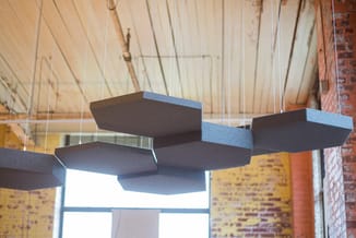 Zwarte hangende akoestische panelen in trapvormige opstelling boven werkplek, tegen bakstenen muur en raampartij.