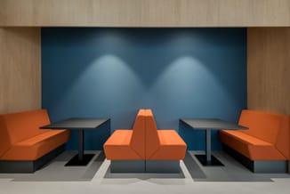 Moderne kantoorlounge met twee oranje banken, centrale oranje poef, twee zwarte tafels en blauwe akoestische wand met spotlig