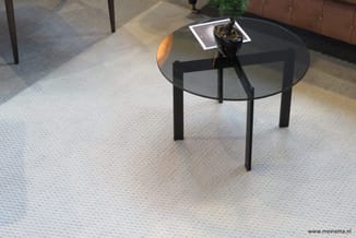 Ronde glazen bijzettafel met zwart metalen onderstel op licht tapijt, met plant en magazine erop in kantoorruimte.