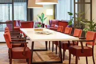 Moderne kantoorruimte met witte vergadertafel, oranje stoelen, rode zitgroep en groene planten onder hanglamp.