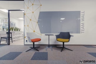 Modern kantoorlounge met twee draaifauteuils (grijs/oranje en grijs/geel) rond zwart tafeltje, gouden netwerk-wandpaneel, bla