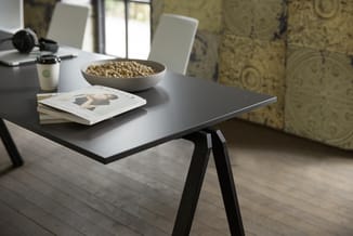 Zwarte bureau met wit blad op zwarte schraagpoten. Tafelblad met stapel boeken, kopje koffie, bowl met noten en koptelefoon.