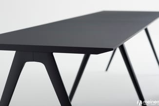 Zwarte vergadertafel met trapeziumvormig blad en minimalistische poten, detail van hoekgeometrie en onderstel.