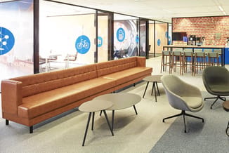 Moderne kantoorlobby met cognackleurige leren bank, grijze stoelen, ronde tafels, en glazen wanden met blauwe fietssymbolen.
