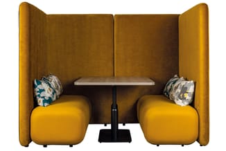 Gele privacy booth met twee zitplaatsen, gevoerde wanden, tafel tussen stoelen, decoratieve kussens.