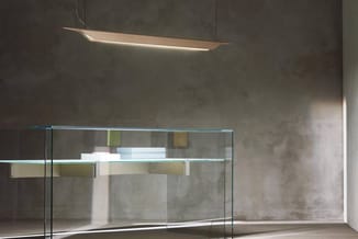 Glazen vergadertafel met turquoise rand, witte objecten erop, in betonnen ruimte met hangende houten designlamp.