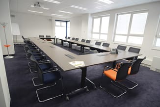 Vergaderzaal met lange zwarte tafel, zwarte en oranje kantooorstoelen, donkerblauwe tapijt, witte wanden en grote ramen.