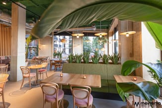 Modern kantoor met houten tafels, roze stoelen met riet, groene plantenbakken, hangende gele lampen en betonnen plafond.