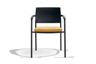 Moderne stoel met zwart metalen frame, zwarte rugleuning en okergele zitting, staand op grijs oppervlak met schaduw.