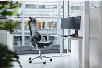 Moderne werkkamer met zwarte ergonomische bureaustoel aan wit bureau met monitor, grote ramen met uitzicht op kantoorgebouwen