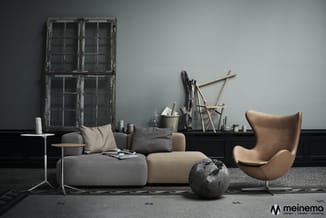 Moderne woonkamer met grijze modulaire bank, cognackleurige leunstoel, houten bijzettafel, vintage raampartij, en decoratieve