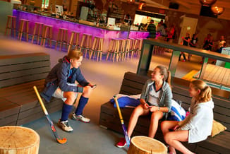 Drie kinderen met hockeysticks zittend op zwarte loungebanken in een moderne sportzaal met paarse verlichting en houten tafel