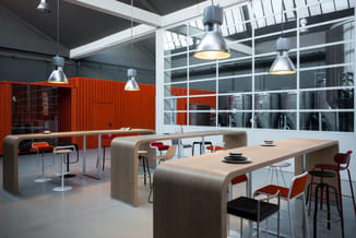 Moderne kantoorruimte met houten vergadertafels, oranje en rode stoelen, hangende metalen lampen, grote ramen en rode contain