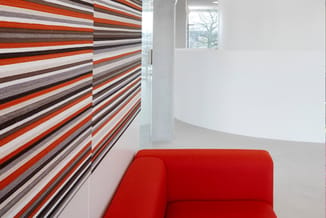 Rode stof sofa tegen witte wand, naast gestreepte wandbekleding in rood, wit, grijs en bruin met diagonaal patroon.