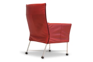 Rode leunstoel met hoge rug, gestoffeerd leer, zilveren metalen frame met wieltjes, tegen witte achtergrond.