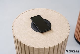 Beige cilindrische poef met verticale groeven en zwarte draadloze oplaadmat met smartphone erop.