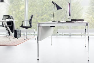 Modern kantoor met wit bureau met metalen onderstel, zwarte bureaustoel, zwarte bureaulamp en monitor. Op achtergrond tweede