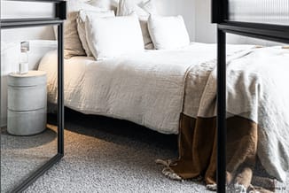 Zwart metalen bed met witte kussens en beige linnen, betonnen nachtkastje met glas, grijze vloerbedekking.