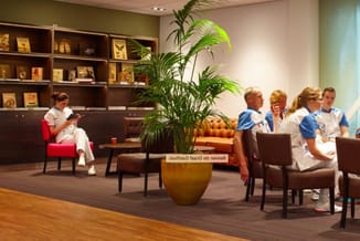 Wachtruimte met bruine fauteuils, rode stoel, houten tafel, gele plantenpot, groene plant, donkere wandkast en meerdere perso