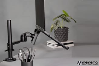 Zwart bureauarrangement met monitorarm, bureaulamp, pennenbakje en plantenpot op witte boeken, tegen grijze wand.