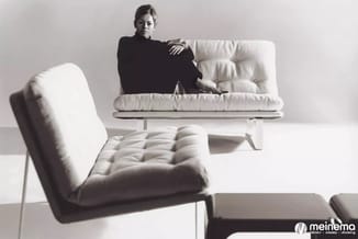 Witte gestoffeerde fauteuil en laag design sofa in minimalistische showroom setting met vrouw in zwarte kleding.
