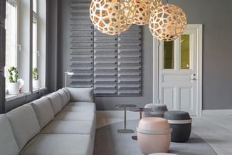 Moderne kantoorruimte met grijze bankstel, houten hanglampjes, raamkozijnen met shutters en ronde poefjes in beige en zwart.