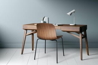 Houten bureau met ladeblokken, houten stoel met metalen poten, witte bureaulamp, pennenbakje en notitieboeken op grijze achte
