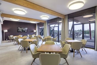 Lounge-ruimte met houten tafels, gele/groene fauteuils, houten balkenplafond, ronde LED-verlichting, bruine accentmuur, grote