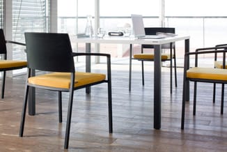 Modern kantoor met witte tafel, zwarte poten en gele stoelen met zwarte ruggen rond een bureau met laptops.