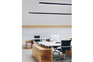 Modern kantoor met houten bureaublokken, witte werkbladen, zwarte bureaustoelen, verlichtingslijnen aan plafond en witte wand