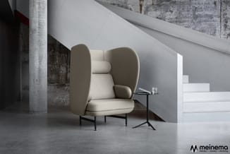 Beige fauteuil met hoge, afgeronde rugvering en zwart metalen onderstel in industriële ruimte met betonnen elementen.