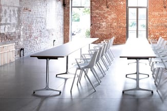 Moderne kantoorruimte met twee zwarte tafels, witte stapelstoelen en bakstenen muren met grote ramen.