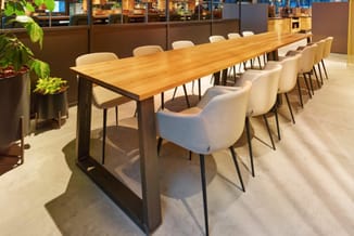 Lange houten vergadertafel met zwart onderstel en witte stoelen in moderne kantoorruimte met groene planten.