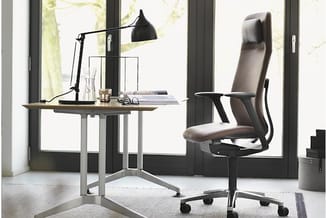 Werkplek met grijs bureaumeubel, zwarte ergonomische bureaustoel met hoofdsteun, zwarte bureaulamp en raam op achtergrond.