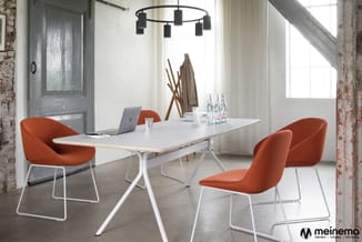 Vergaderruimte met witte rechthoekige tafel, vier oranje stoelen met metalen frame, zwarte hanglamp, grote ramen en industrië