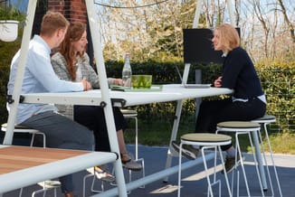 Drie personen bij een witte vergadertafel in een moderne buitenruimte met beige stoelen, monitor en laptops.