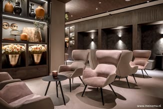 Moderne lounge met roze fluwelen fauteuils rond zwart tafeltje, verlichte display kast met decoratie en bloemen, donkere hout