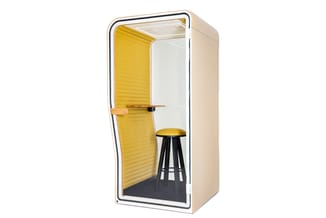 Akoestische belcel met gele wanden, houten deur, zwart barkriukje en grijze vloer tegen beige buitenwanden.