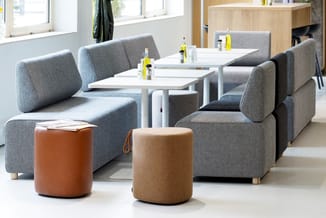 Lounge-opstelling met grijze modulaire banken, witte tafels, bruine en beige poefzitjes, glazen flessen en grote ramen in mod