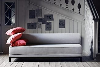 Grijze loungebank met rode kussens in minimalistische ruimte met houten vloer en wanddecoratie.