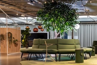 Groene loungebank met zwarte poten in showroom, hangend bladplant, houten panelen, oranje hangende lampen, lichte vloer.