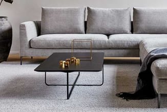 Grijze hoekbank met grijze kussens, zwarte salontafel met metalen onderstel en gouden decoratieve objecten op beige vloerklee