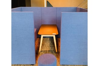 Akoestische belcel met blauwe wanden, oranje tafelblad en houten poten in kantoorruimte met gestreepte vloer.