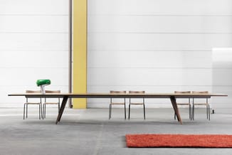 Lange vergadertafel met donker houten blad en schuine poten, bezet met stoelen, in industriële ruimte met gele kolom en oranj