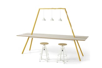 Geel stalen trapeziumvormig frame met lichte houten werkblad, drie hangende witte metalen lampen en twee witte verstelbare kr